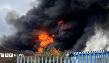 An explosion of orange flames rises into a billowing plume of black smoke above the steel fencing and brown brickwork of the industrial estate.