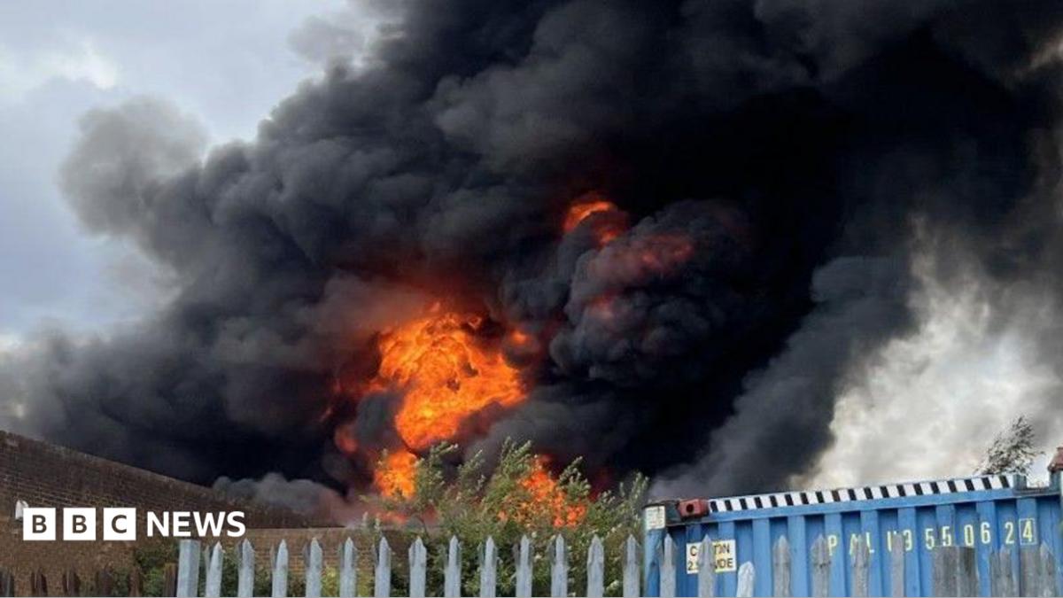An explosion of orange flames rises into a billowing plume of black smoke above the steel fencing and brown brickwork of the industrial estate.