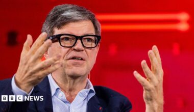 Yann LeCun wears fashionable thick-rimmed glasses, a light blue shirt and navy jacket. He is using his hands to articulate a point while speaking at a conference.