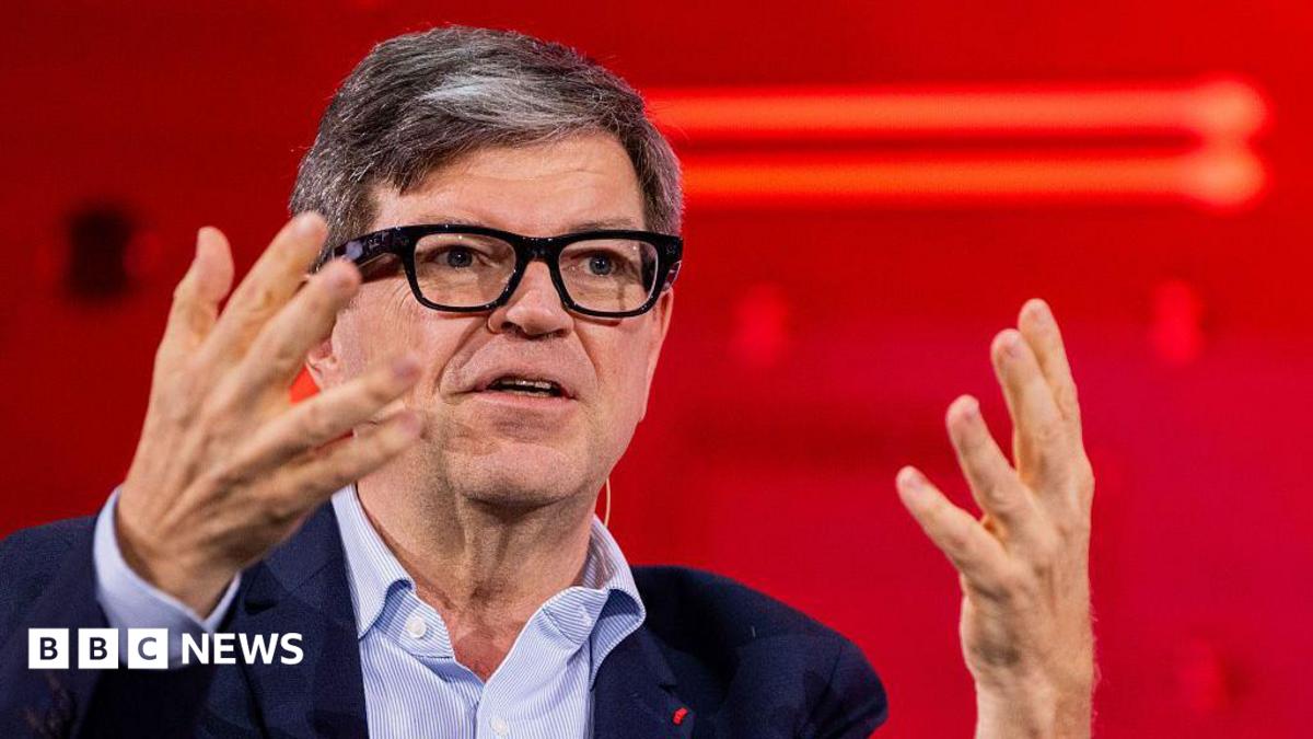 Yann LeCun wears fashionable thick-rimmed glasses, a light blue shirt and navy jacket. He is using his hands to articulate a point while speaking at a conference.