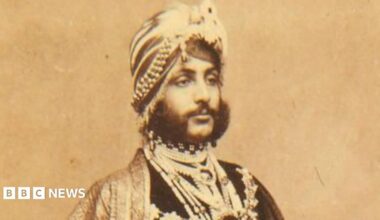 A portrait of Duleep Singh on paper that has aged over time and gone brown. He looks away from the camera and is expressionless. He has 19th Century traditional Indian headwear on as well as clothing. He also has a dark and thick beard.