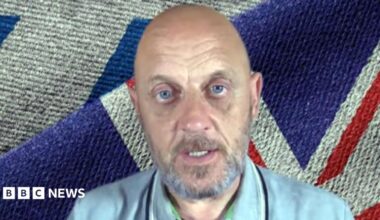 A bald and bearded man with blue eyes sitting in front of a Union flag