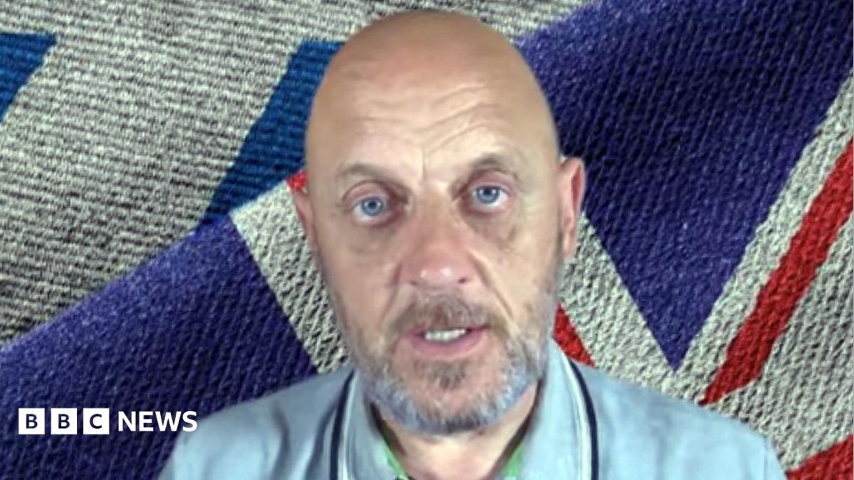 A bald and bearded man with blue eyes sitting in front of a Union flag