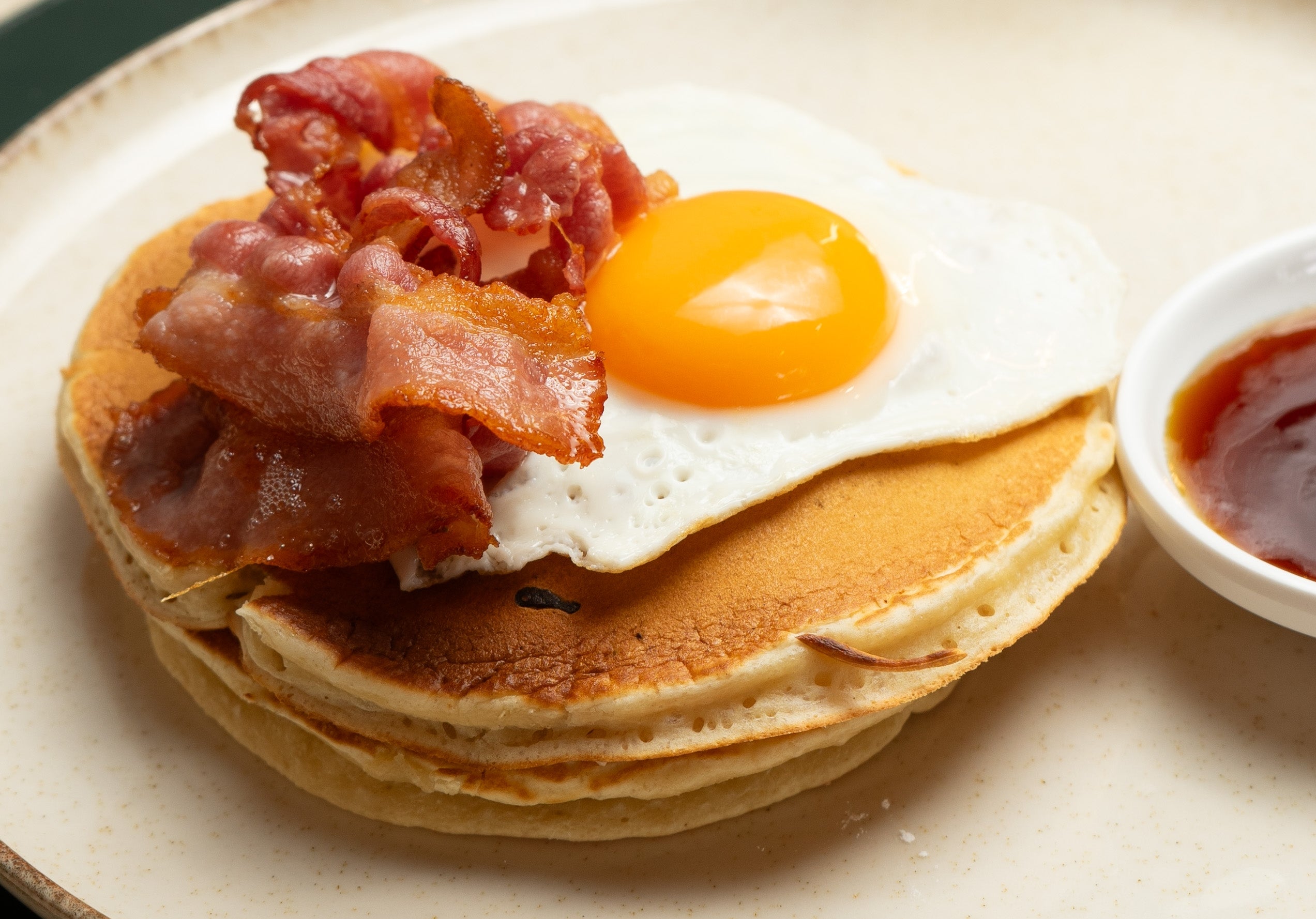 Stack of pancakes topped with fried egg and crispy bacon, served with a side of syrup, on a table with a glass of orange juice