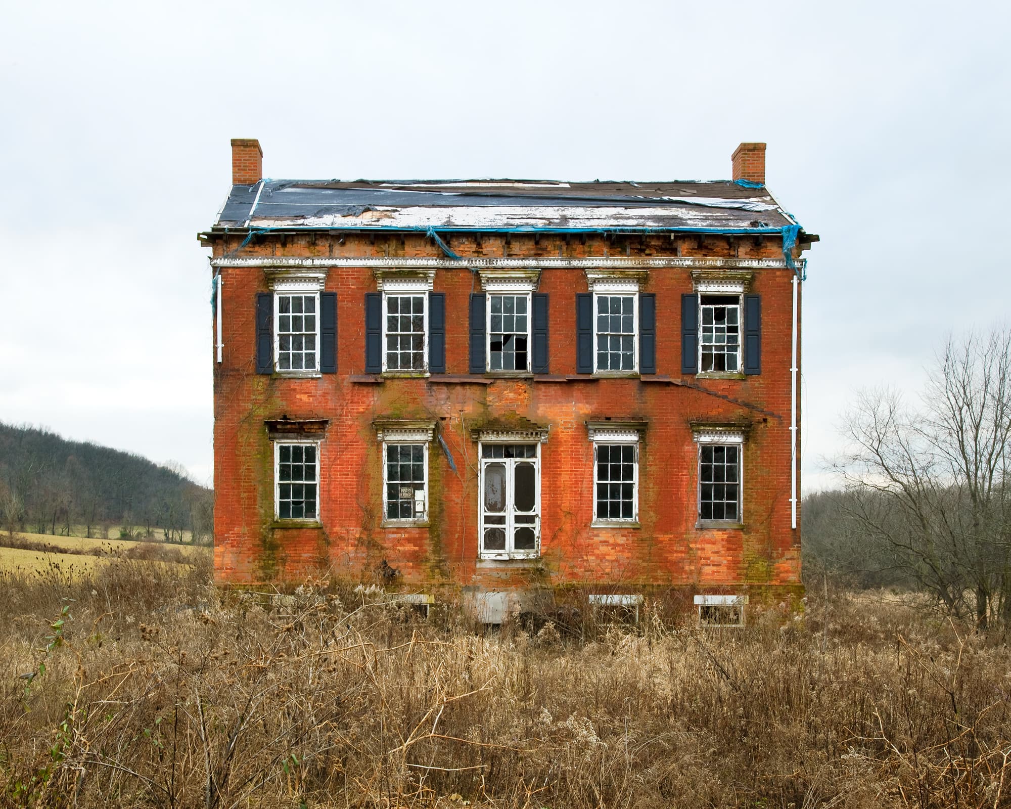 A grand, decaying two-story red brick historic mansion with broken windows and a tarp-covered roof sits abandoned amidst an overgrown field of tall, dry brush.