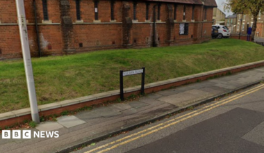 A street view image of a road with double yellow line and a pavement. A street name sign is visible with a grass bank behind it and a red brick building.