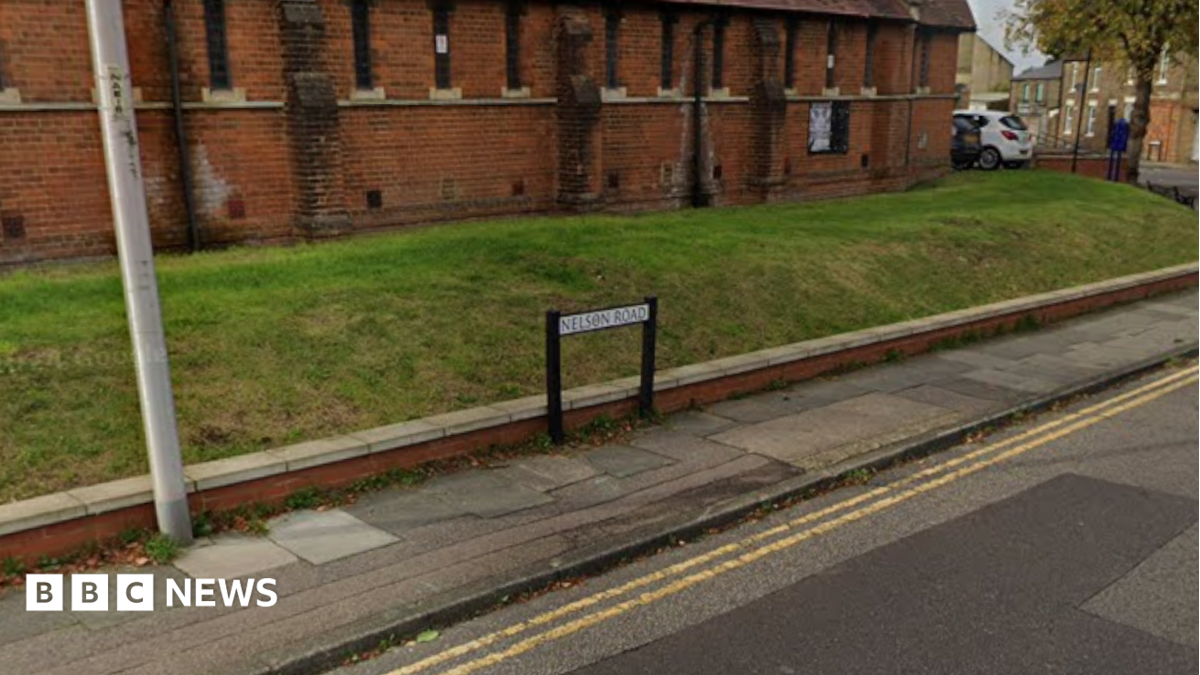 A street view image of a road with double yellow line and a pavement. A street name sign is visible with a grass bank behind it and a red brick building.