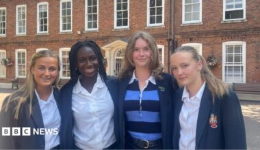 Four girls wearing blue school blazers stand in a row in front of a large brick building with large white frame windows.