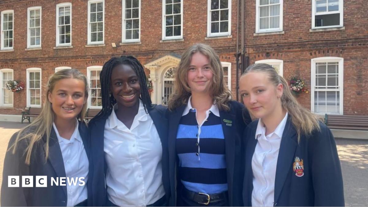 Four girls wearing blue school blazers stand in a row in front of a large brick building with large white frame windows.