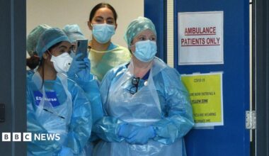 Four medics are standing at the doors of an emergency department in the pandemic. They are wearing plastic blue gowns and have face masks on. One has a sign on her chest saying "ED Nurse".