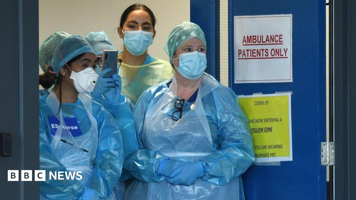 Four medics are standing at the doors of an emergency department in the pandemic. They are wearing plastic blue gowns and have face masks on. One has a sign on her chest saying "ED Nurse".