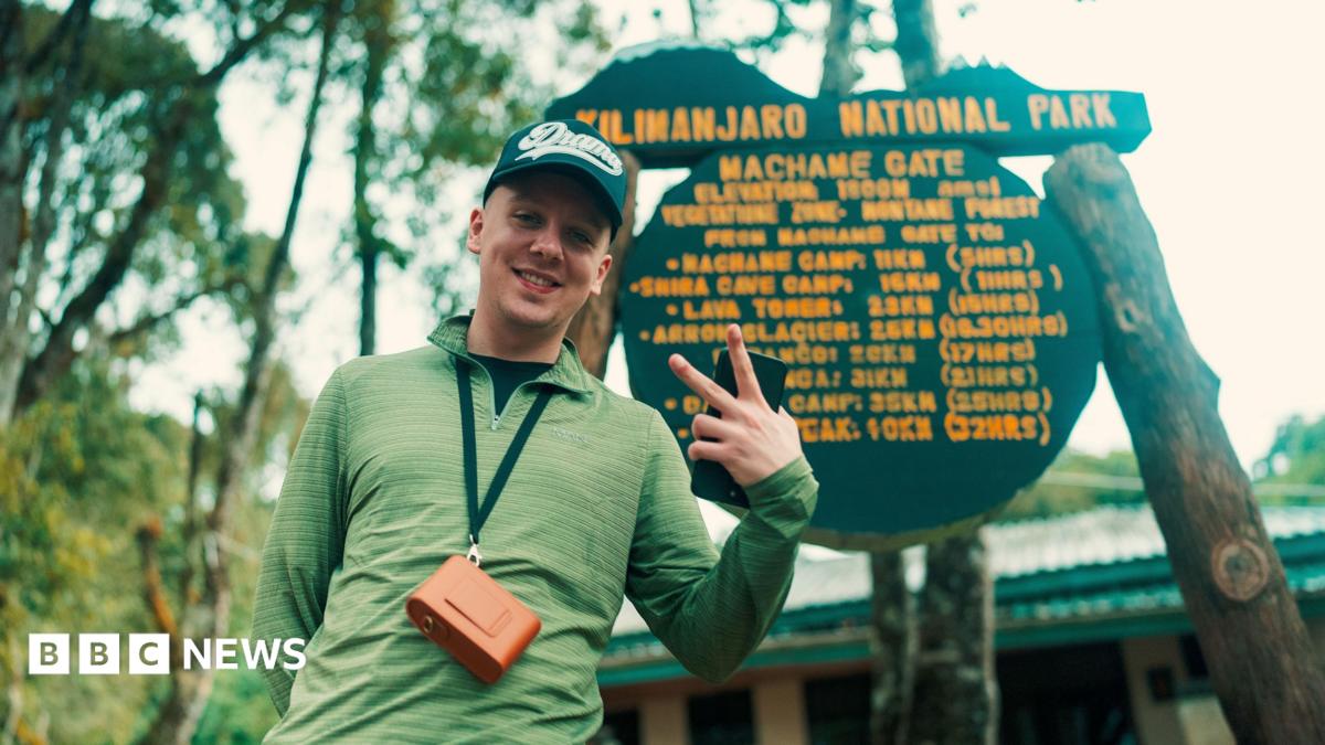 Aitch posts with his sister Gracie, who is kissing him on the cheek. Aitch is wearing black hoodie with the hood up. Gracie is wearing a pink top.