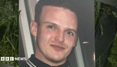 A large print out of a photo of Stephen Morrisson is propped up against foliage. He is wearing a black polo shirt with white stripes on the collar. He has a slight smile and has dark brown hair.