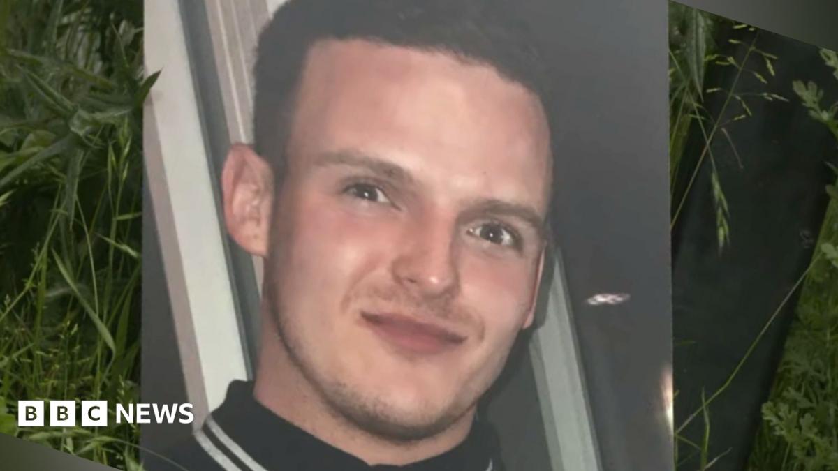 A large print out of a photo of Stephen Morrisson is propped up against foliage. He is wearing a black polo shirt with white stripes on the collar. He has a slight smile and has dark brown hair.
