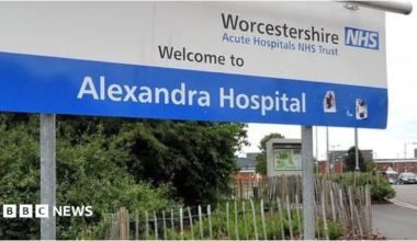 A blue and white hospital sign on a roadside, which reads Worcestershire Acute Hospital NHS Trust Welcome to Alexandra Hospital