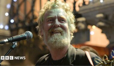 Glen Hansard has curly reddish/grey hair and beard and is singing while playing a guitar