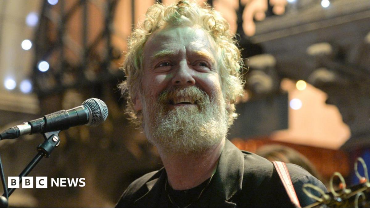 Glen Hansard has curly reddish/grey hair and beard and is singing while playing a guitar