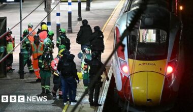 Black-clad police officers and emergency crews wearing green-and-yellow coveralls and helmets on the platform of Huntingdon railway station, where a train has stopped.