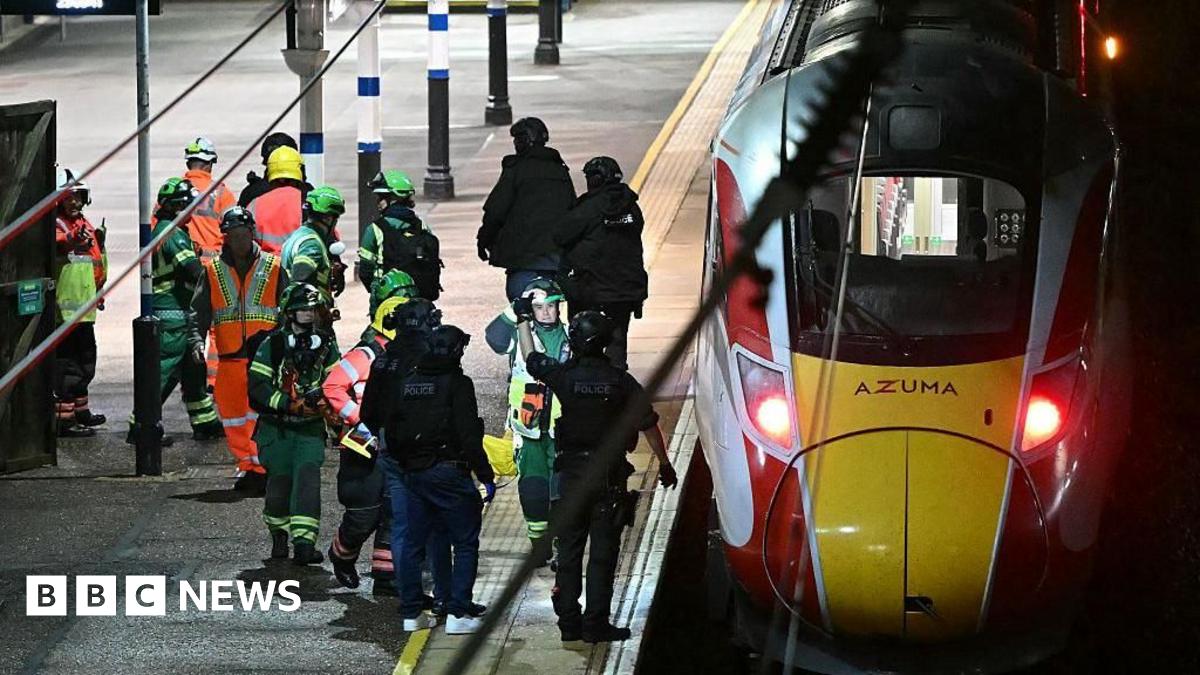 Black-clad police officers and emergency crews wearing green-and-yellow coveralls and helmets on the platform of Huntingdon railway station, where a train has stopped.