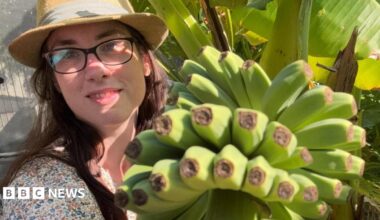 A woman with glasses and a hat posing next to a bunch of bananas