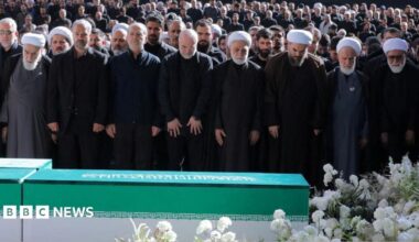 Iranian President Masoud Pezeshkian, Iran's Parliament speaker Mohammad Baqer Qalibaf and Chief Justice of Iran Gholam-hossein Mohseni Ejei with officials attend a prayer during a public farewell ceremony to pay their respects to late Supreme Leader Ayatollah Ali Khamenei. A large crowd can be seen in the background as everyone stands looking at the coffin