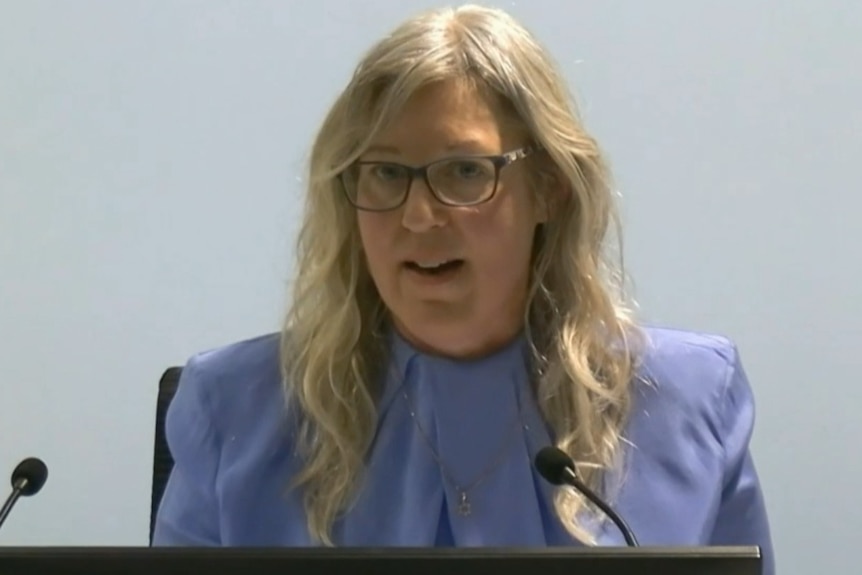 A blonde woman with shoulder length hair wearing black framed glasses and a blue shirt.