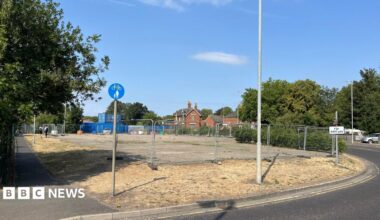 The derelict land where the coffee shop will be built. It has concrete paving surrounded by grass and with metal fencing around the site