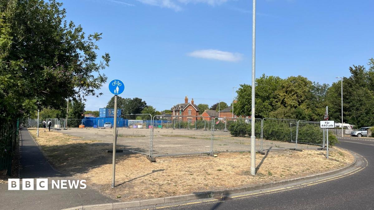 The derelict land where the coffee shop will be built. It has concrete paving surrounded by grass and with metal fencing around the site