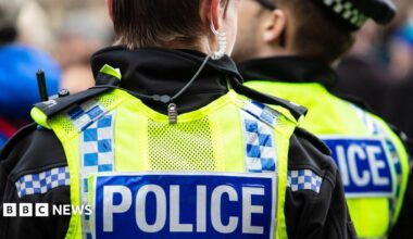 Two police officers wearing yellow hi-vis vests with "police" written on the back. One officer has an earpiece. They both have their backs to the camera.