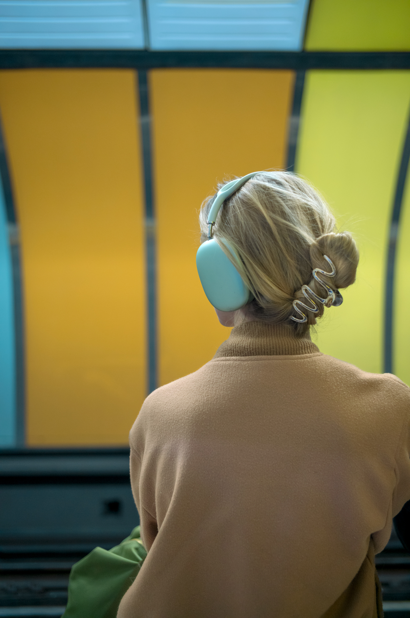 Seen from behind, a woman with teal headphones and a hair clip stands before a stained-glass-like wall of yellow, teal and green panels.
