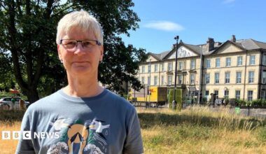 A man with white hair standing in a wildflower meadow. He is wearing glasses and a blue T-shirt with birds on it. Behind him stands a large, three-storey, Victorian-style building in yellow brick.