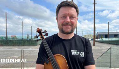 Dónal is holding a fiddle and smiling. He's wearing a black top that says Belfast TradFest 2026. The Titanic slipways are in the background, with metal poles lining the space. It's a cloudy day.