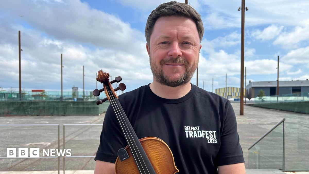 Dónal is holding a fiddle and smiling. He's wearing a black top that says Belfast TradFest 2026. The Titanic slipways are in the background, with metal poles lining the space. It's a cloudy day.