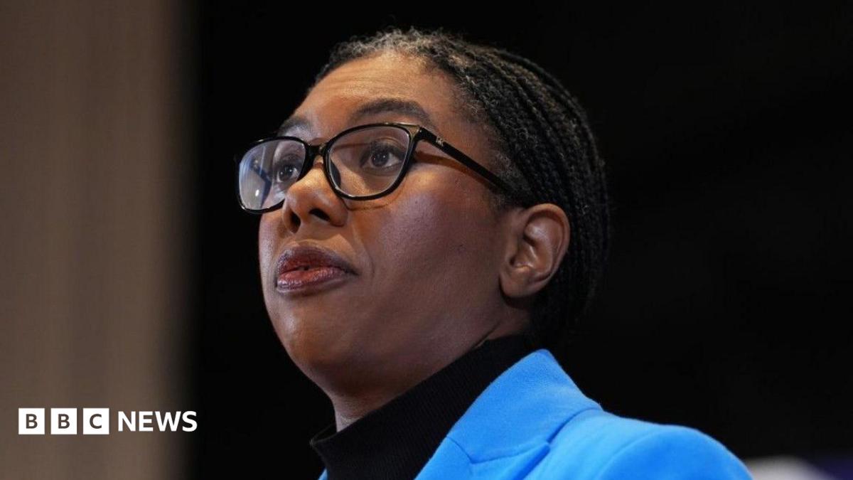 Kemi Badenoch has a serious expression and is wearing a sky blue jacket over a black turtleneck top. She has her braided hair tied back in a low ponytail and wears black spectacles.