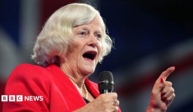 Close-up of Ann Widdecombe, wearing a red blazer, speaking into a microphone, with her hand raised.