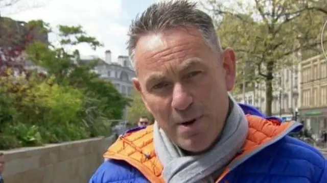 Marcus Farmer has brown hair with grey temples and wears a blue anorak with orange lining and a grey scarf.
