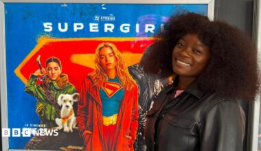 A woman in a black leather jacket is smiling at the camera. She is standing next to a Supergirl film poster