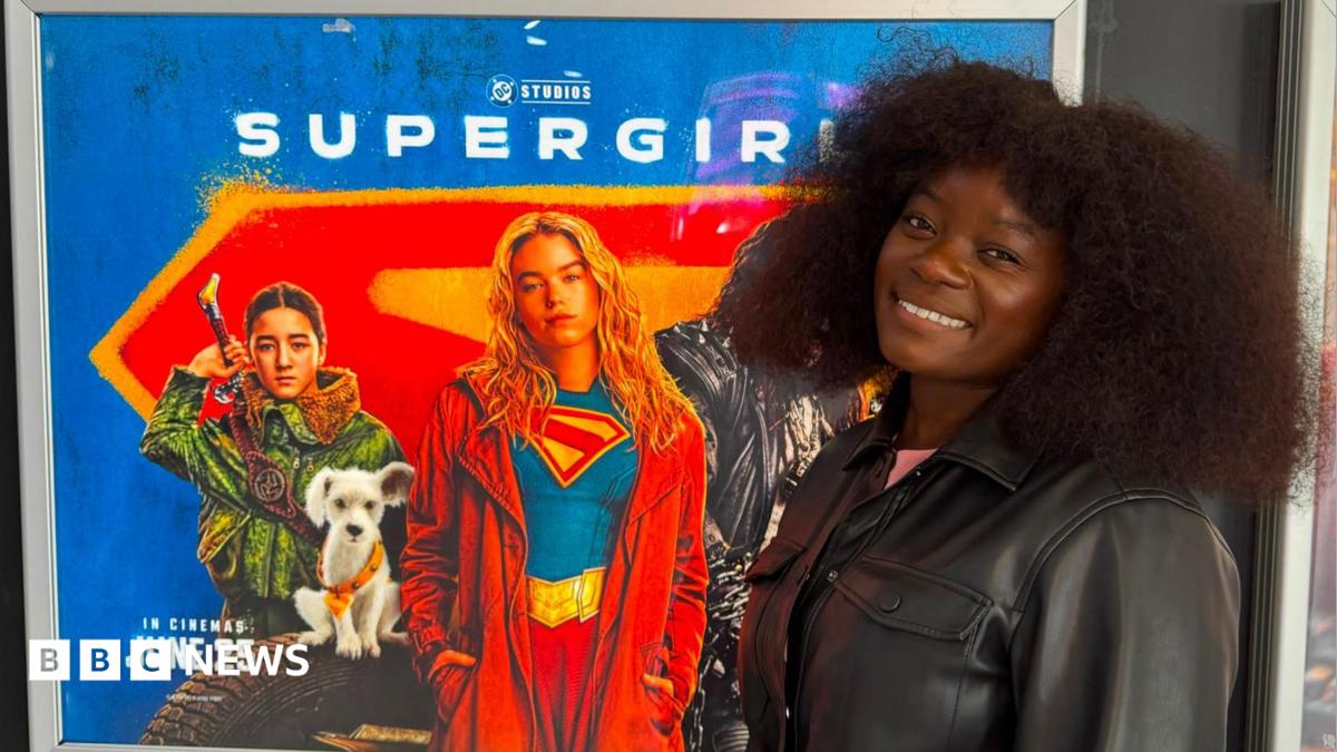 A woman in a black leather jacket is smiling at the camera. She is standing next to a Supergirl film poster