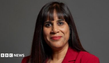 The MP Sureena Brackenridge is looking directly at the camera. The background is grey. She has dark long hair and is wearing a red jacket.