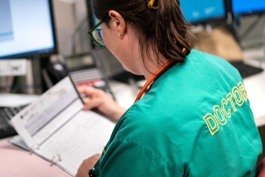 A dark-haired woman in scrubs refers to a documeent while sitting at a desk.