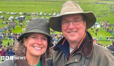 Alice Dearing and her husband John stood next to each other having their picture taken outside. They are dressed in coats and hats each and are looking directly at the camera and smiling. Alice is on the left and John is on the right. They are at an event with crowds of people in the background.
