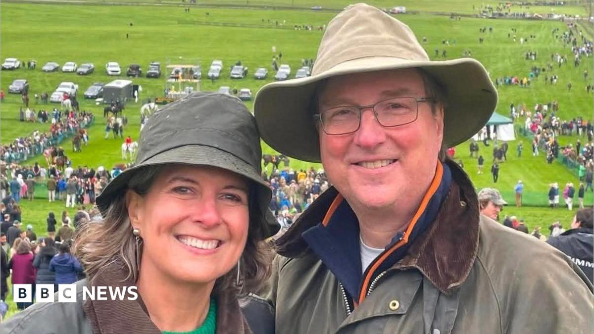 Alice Dearing and her husband John stood next to each other having their picture taken outside. They are dressed in coats and hats each and are looking directly at the camera and smiling. Alice is on the left and John is on the right. They are at an event with crowds of people in the background.