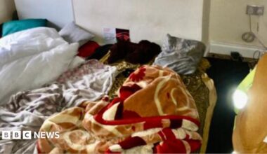 A sparsely furnished room with bedding spread directly on the floor. Several blankets and sheets are piled on patterned floor coverings beneath a chimney breast or wall recess marked by dark damp or mould stains.