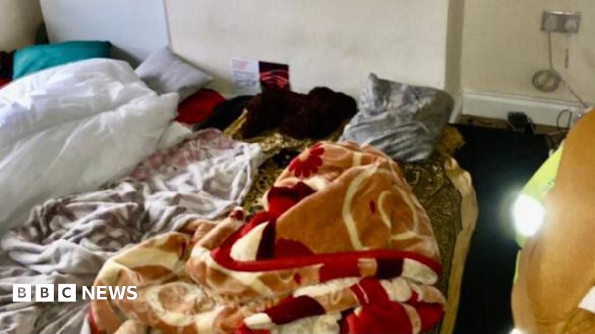 A sparsely furnished room with bedding spread directly on the floor. Several blankets and sheets are piled on patterned floor coverings beneath a chimney breast or wall recess marked by dark damp or mould stains.