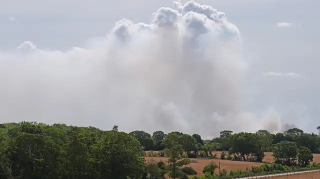 A large smoke plume in the sky above fields and trees.