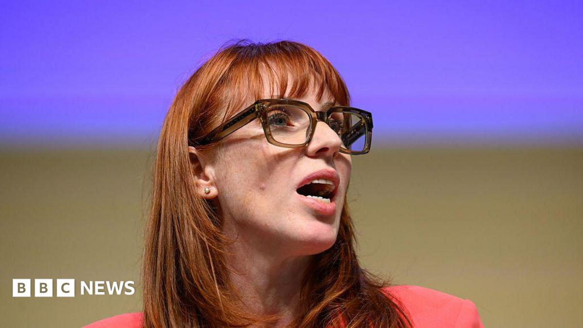 A woman with long, ginger hair and a fringe talks to someone off-camera. She wears thick-rimmed glasses