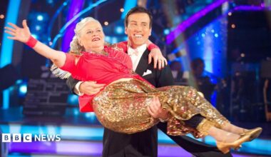 Ann Widdecombe wearing a red top and a golden glittery skirt, being held by Anton Du Beke in a tuxedo