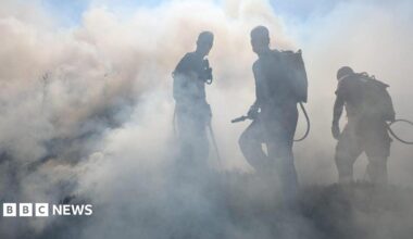 Firefighters wearing backpack water sprayers work through thick smoke while tackling a vegetation fire on open moorland. The figures are silhouetted against dense grey and white smoke that fills most of the scene.
