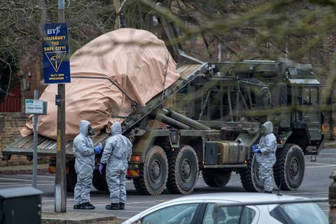 On a street by a sign that says Salisbury the Safe City, people dressed in grey hazmat suits and masks load a covered car onto the back of a military flatbed truck.