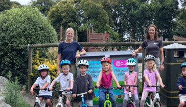A row of children about age four and five on bikes, two women standing behind, at a playground. Trees and blue skies seen behind.